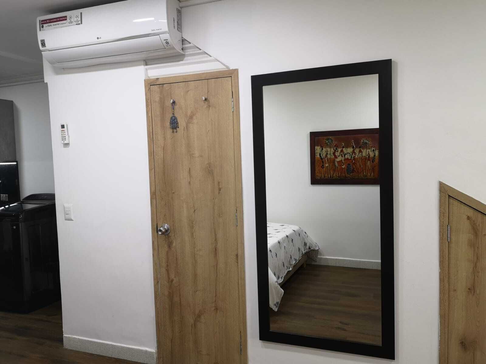 Ein Zimmer mit Holztür, einem Bett mit weißer Bettdecke, einem großen Spiegel und einer Klimaanlage.
