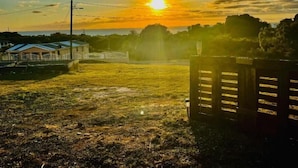 Paisaje rural con un atardecer, campos abiertos, una valla de madera y una pequeña casa con techo azul.