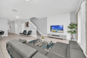 A modern living room with a large TV, grey sofas, a glass coffee table, and a staircase leading to another level.