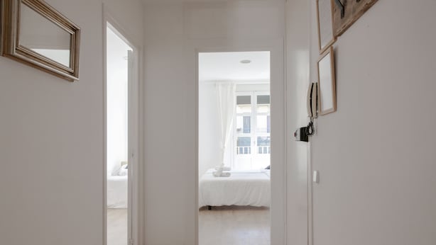 Een gang die leidt naar een slaapkamer met een bed, een houten bankje en een spiegel aan de muur.