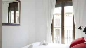 Een hotelkamer met een bed, witte gordijnen, een raam met uitzicht op gebouwen en een zwarte lamp aan de rechterkant.
