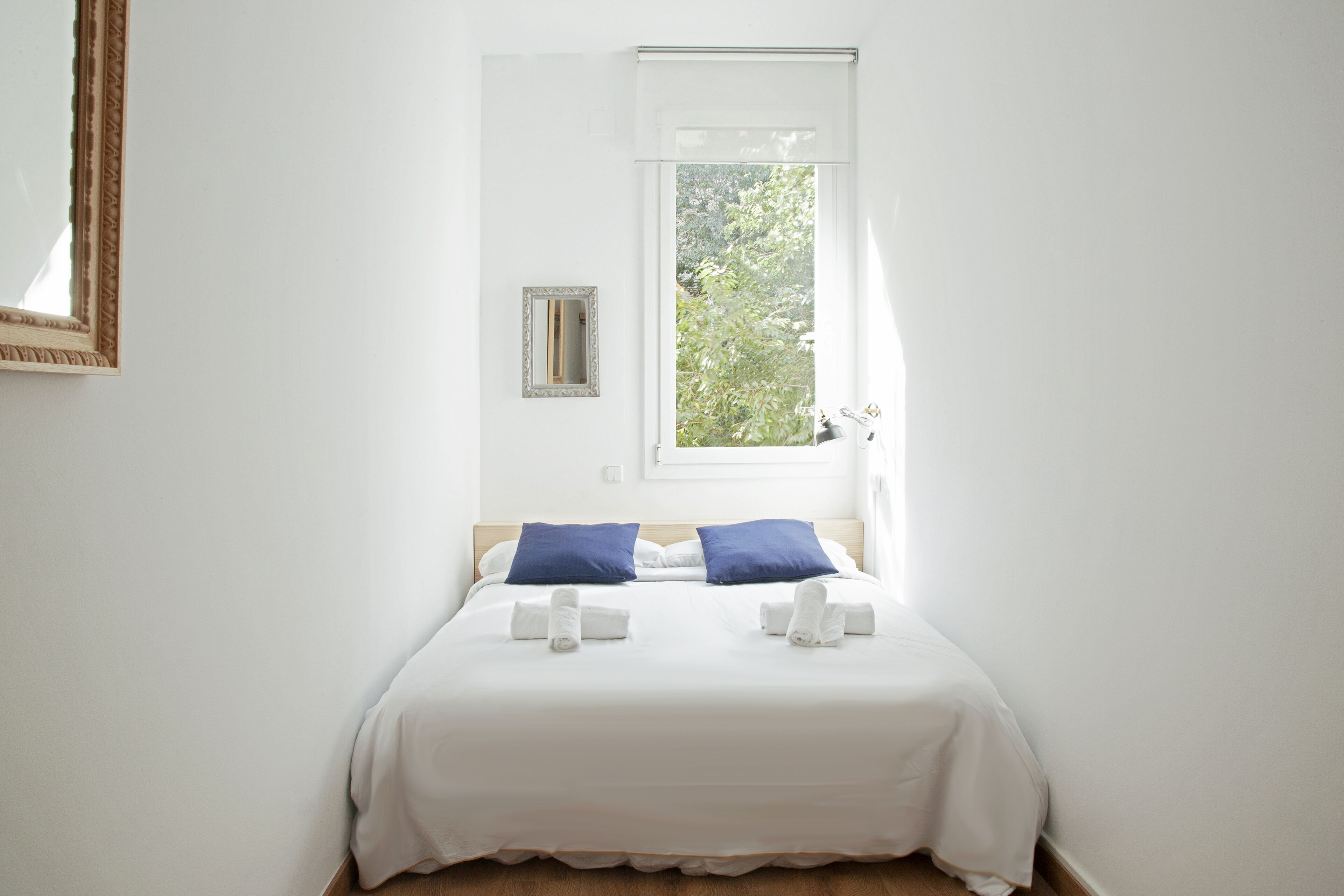 Una cama bien hecha con ropa blanca y dos almohadas azules, un espejo pequeño en la pared y una ventana con vistas al verdor exterior.