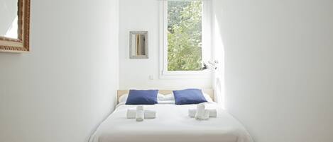 A neatly made bed with white bedding and two blue pillows, a small mirror on the wall, and a window with a view of greenery outside.