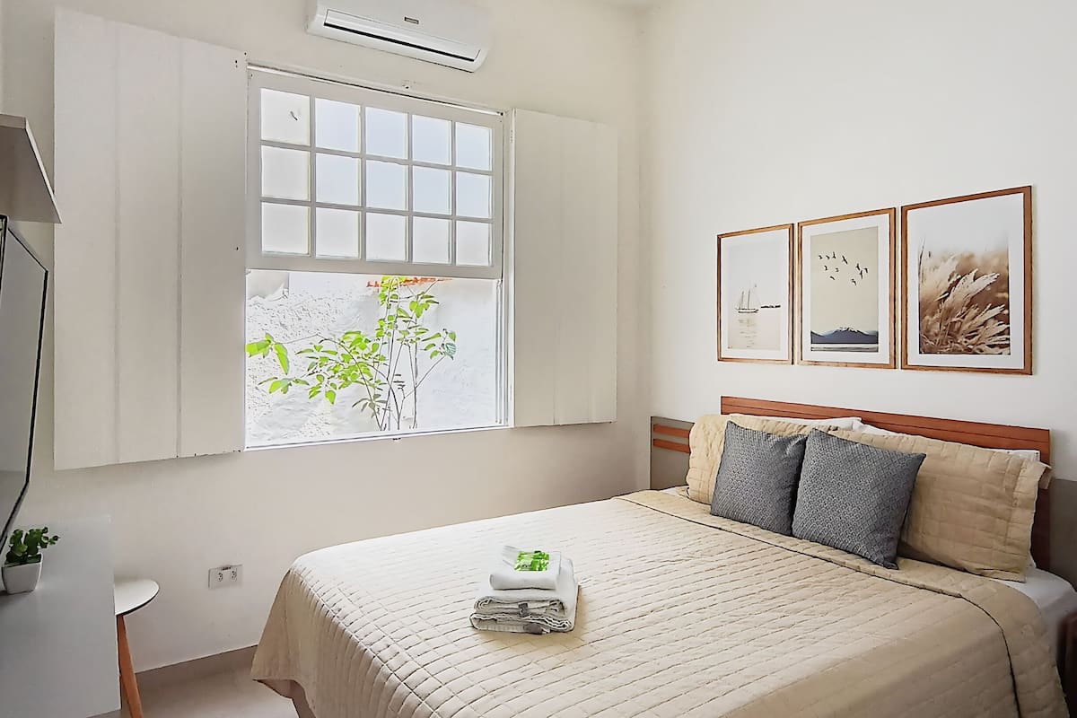 Een slaapkamer met een bed, een raam met zonwering, een wandairconditioner, een nachtkastje en een kleine kamerplant.