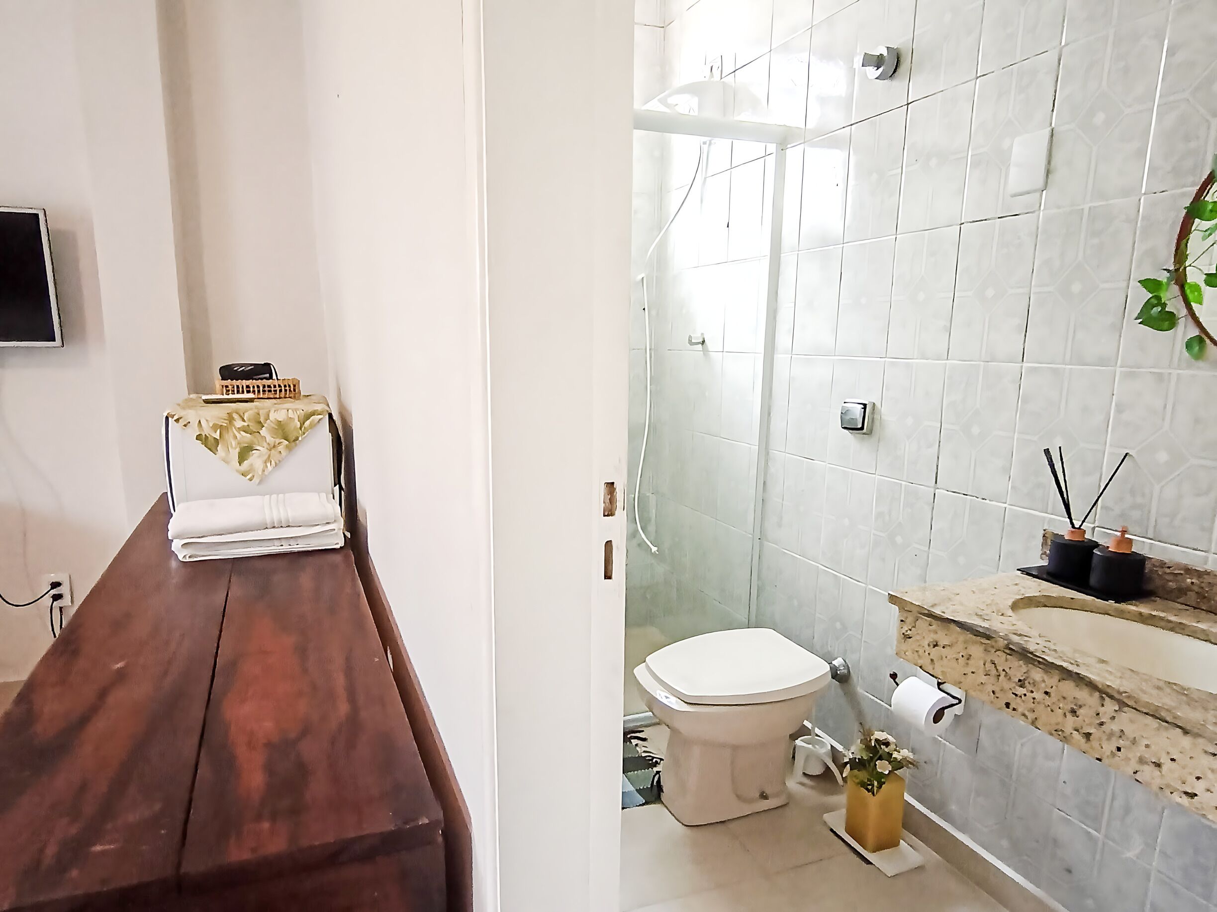 A bathroom with a wooden bench, a toilet, a shower, and a sink with a plant.