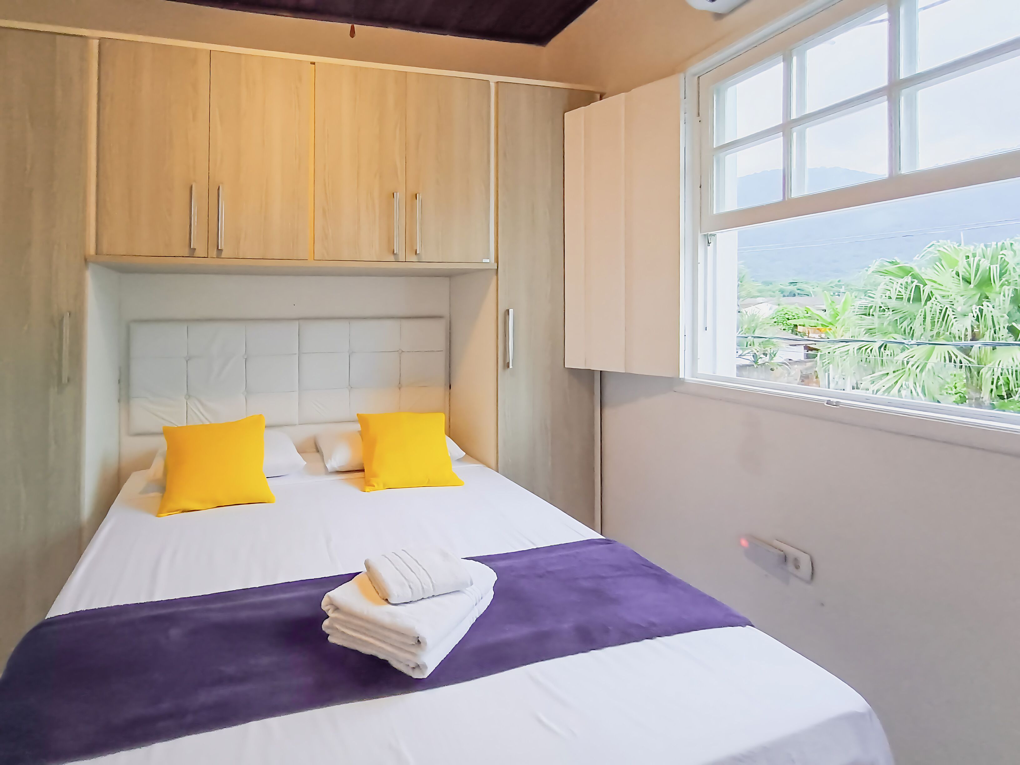 A bed with white bedding and yellow pillows, a purple accent, and a view of greenery through a window.