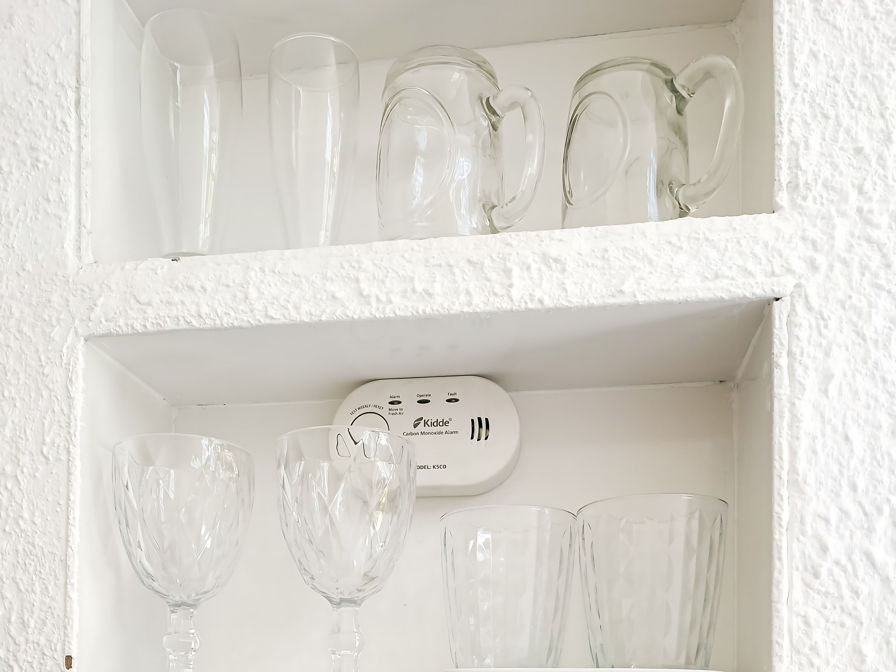 Ein weißes Regal mit klarem Glasgeschirr, darunter Weingläser und ein Kohlenmonoxidmelder.
