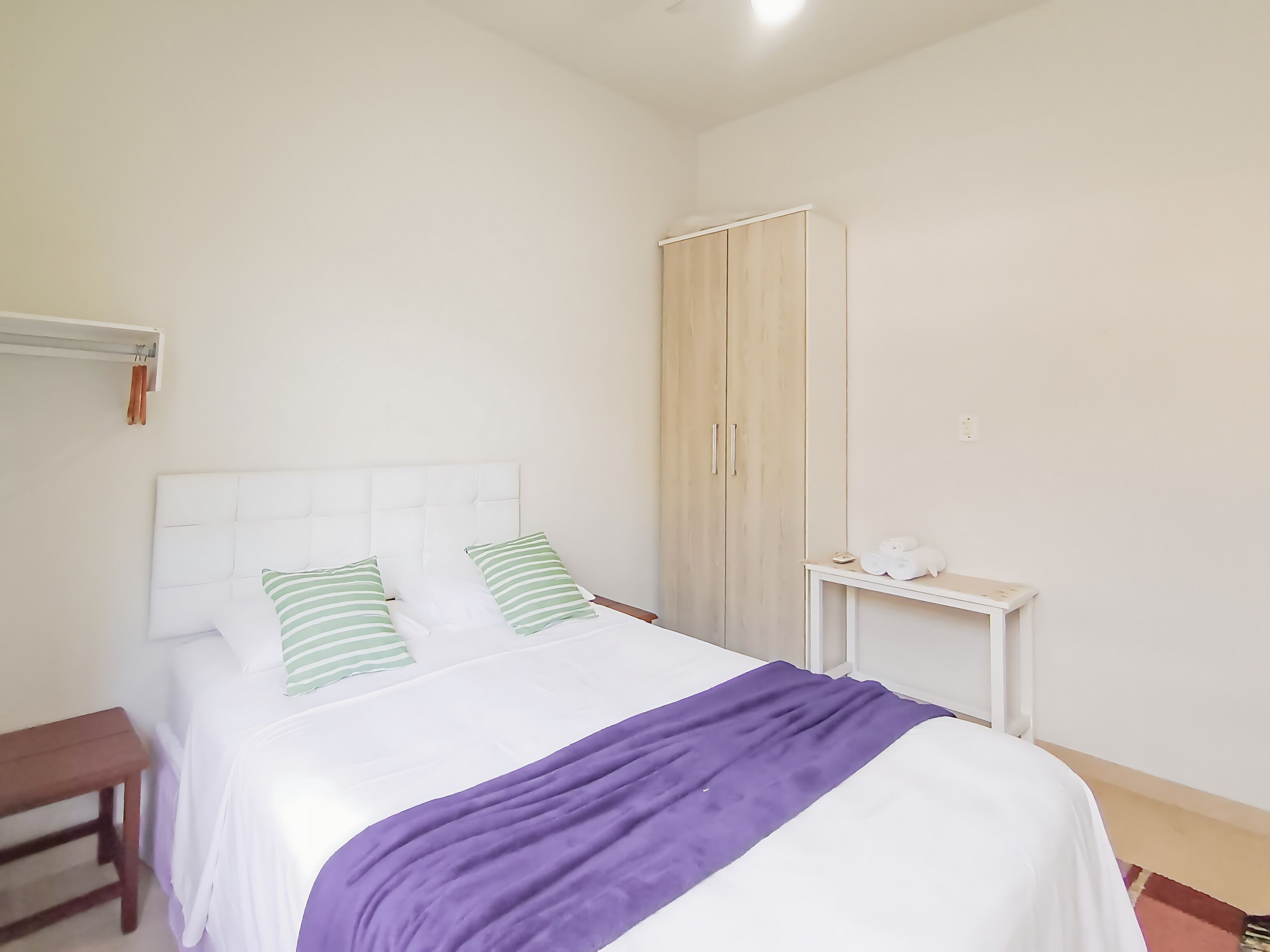 A bed with a white headboard, green striped pillows, a purple blanket, a wooden nightstand, and a wardrobe.