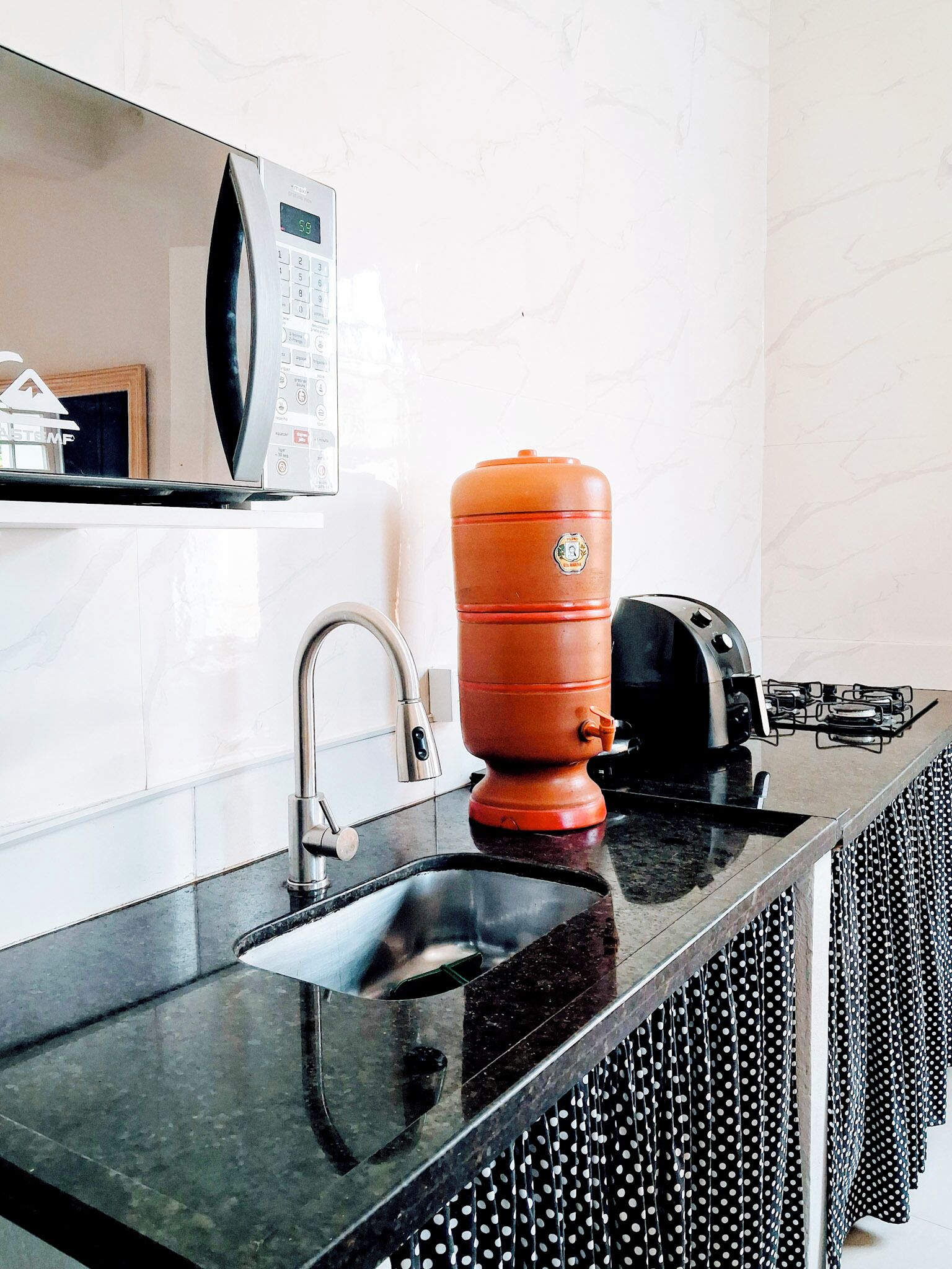 Una cocina con encimera negra, un lavamanos de acero inoxidable, un microondas y un calentador de agua rojo.
