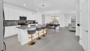 A modern kitchen with white cabinets, a central island with bar stools, and a dining area with a sofa and table. - Stunning 6 Bed Pool Home with Game Room-8964PTD (Davenport)