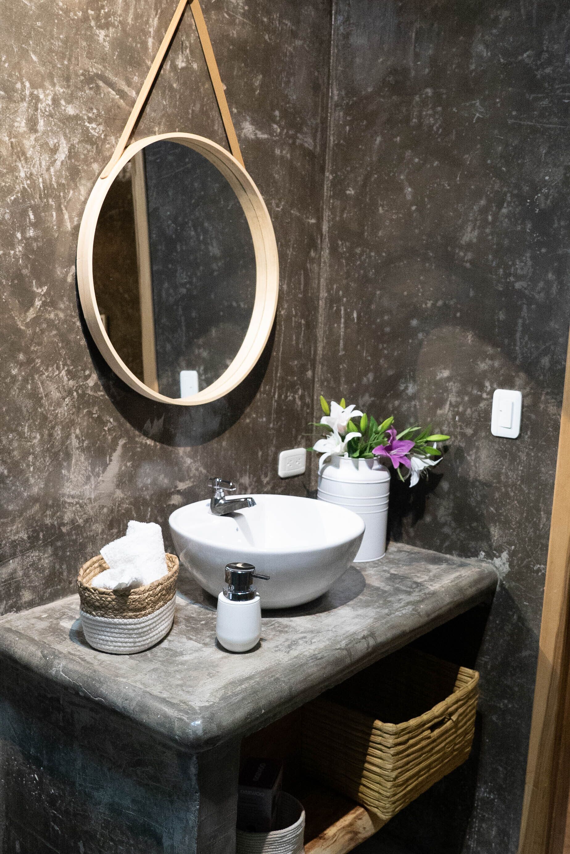 Baño con espejo redondo, lavamanos blanco y un florero con flores.