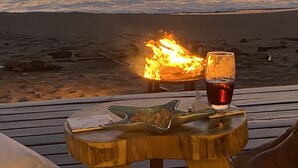 Una mesa de madera con un fuego, un vaso de bebida roja y un vaso de bebida verde en una playa con vista al océano al atardecer.