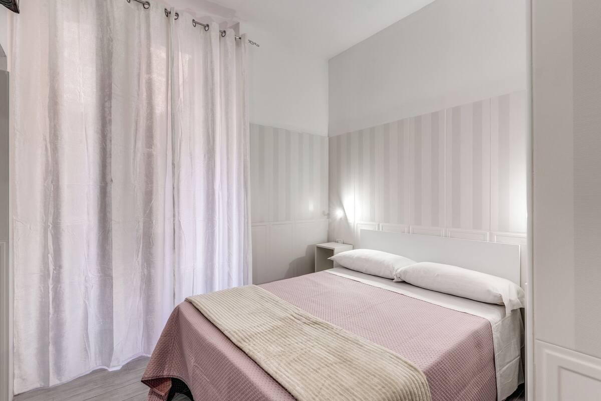 Un dormitorio con cama, cortinas blancas, una mesita de noche y una mesita auxiliar.