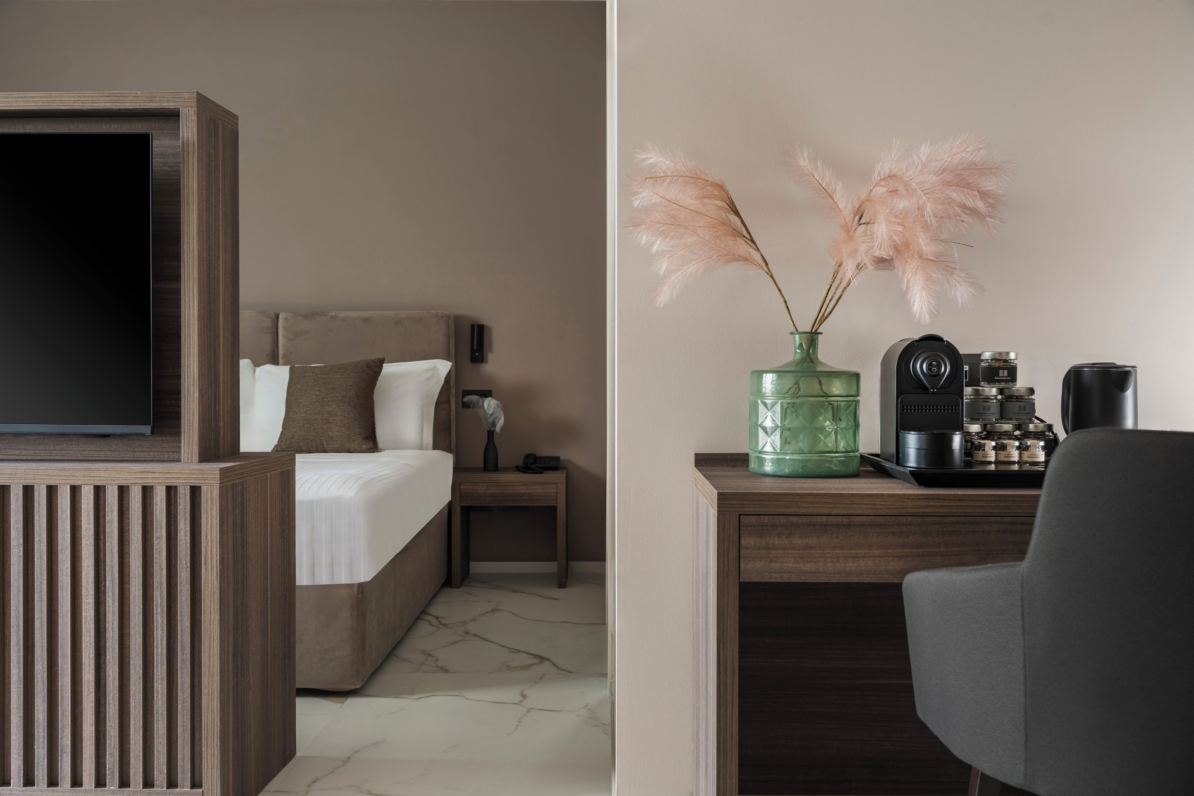 Una habitación de hotel con cama, mesita de noche, escritorio con cámara, jarrón verde con plumas de miscanthus y cafetera.