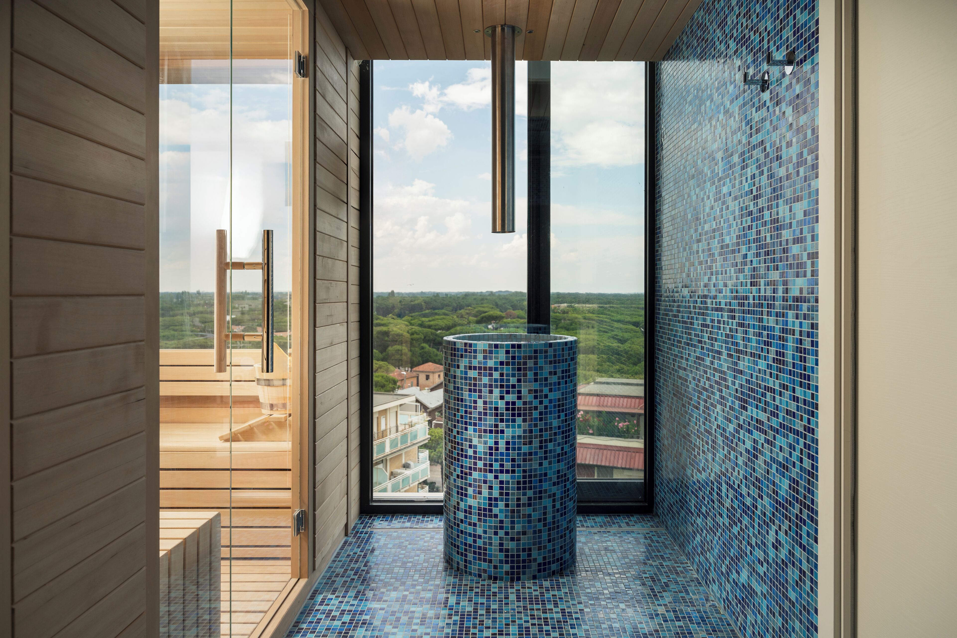 A modern bathroom with a mosaic tile wall, a cylindrical sink, and a view of a balcony and greenery outside.