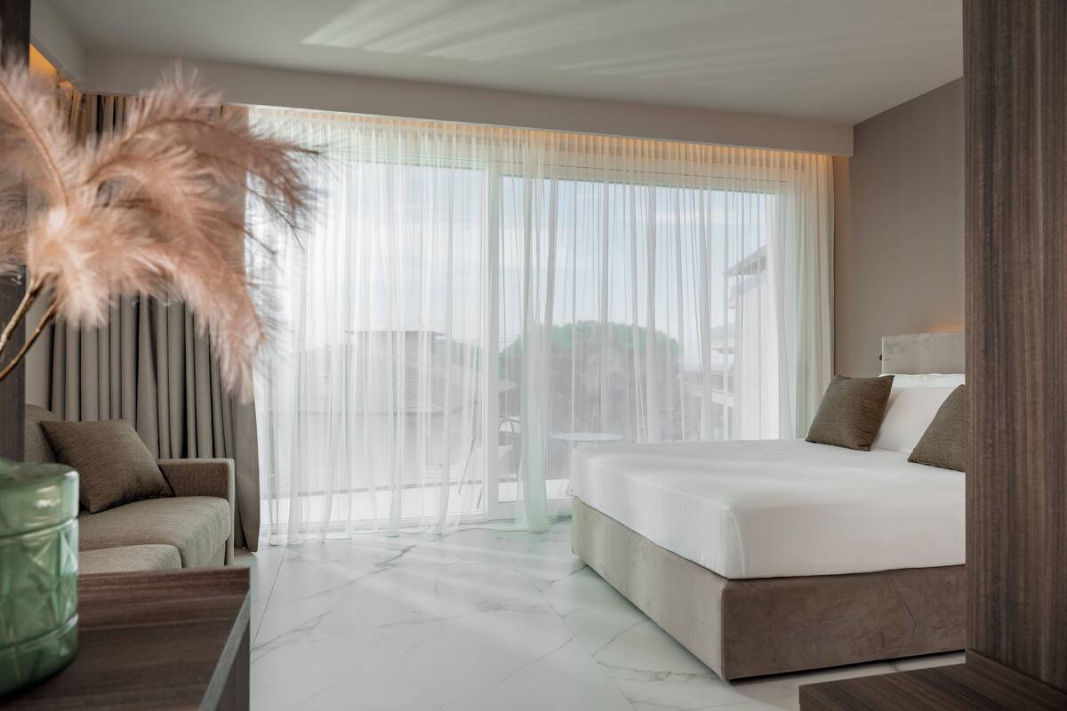 Ein modernes Hotelzimmer mit einem großen Bett, einer Couch und Blick durch transparente Vorhänge.