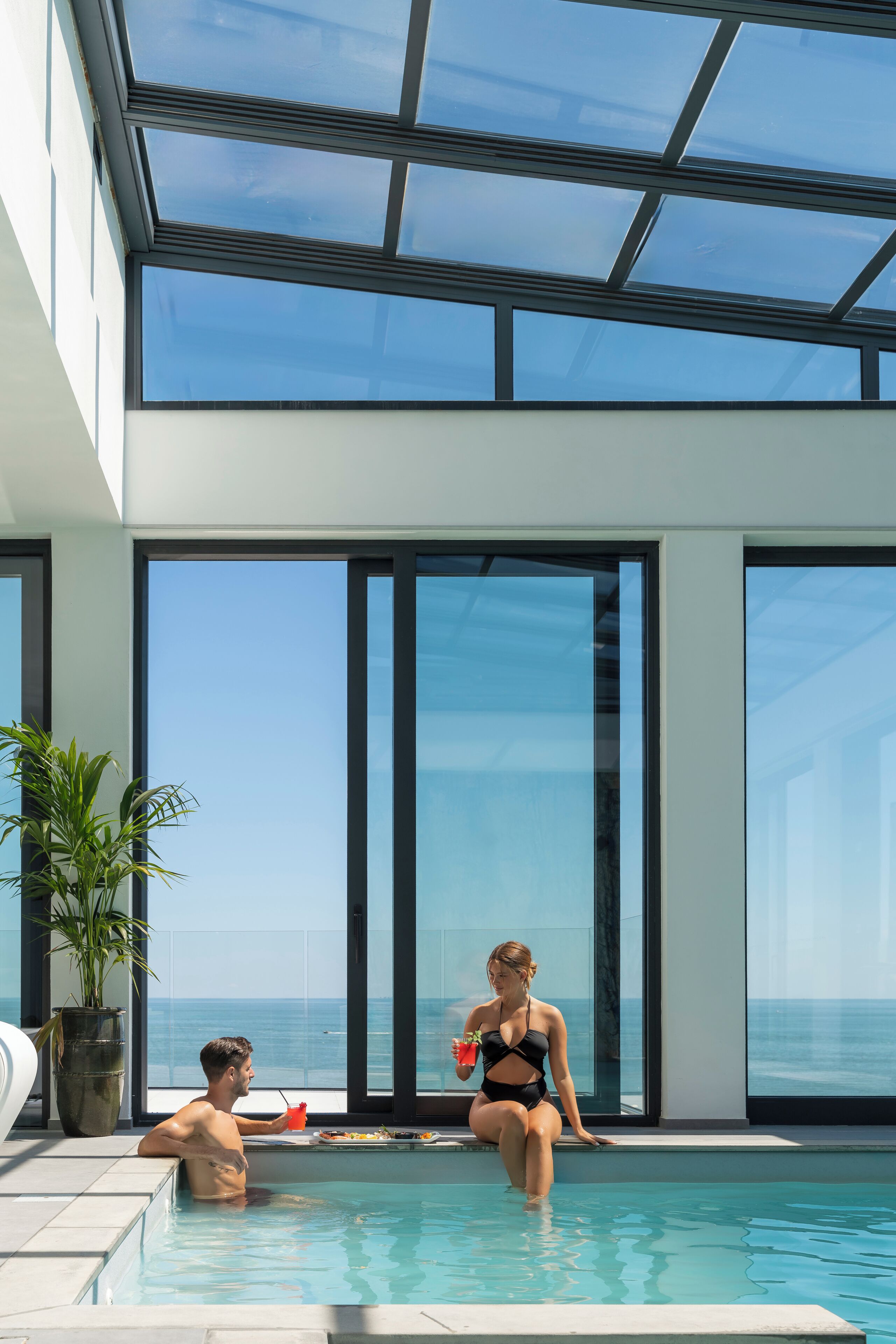A modern indoor swimming pool with large glass windows and a view of the ocean.