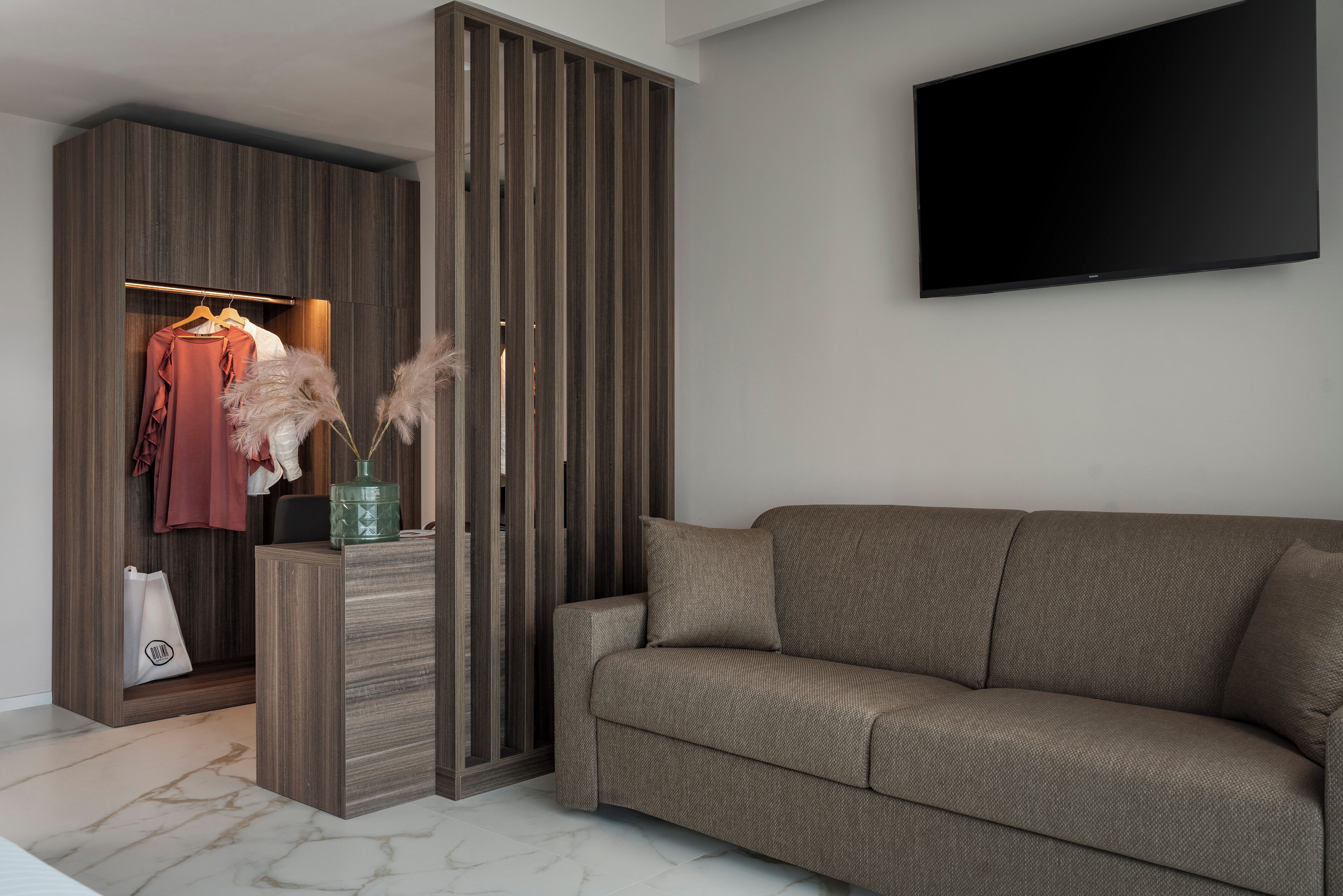 Una sala moderna con un sofá, un televisor montado en la pared, un armario de madera y un perchero con una prenda rosa.