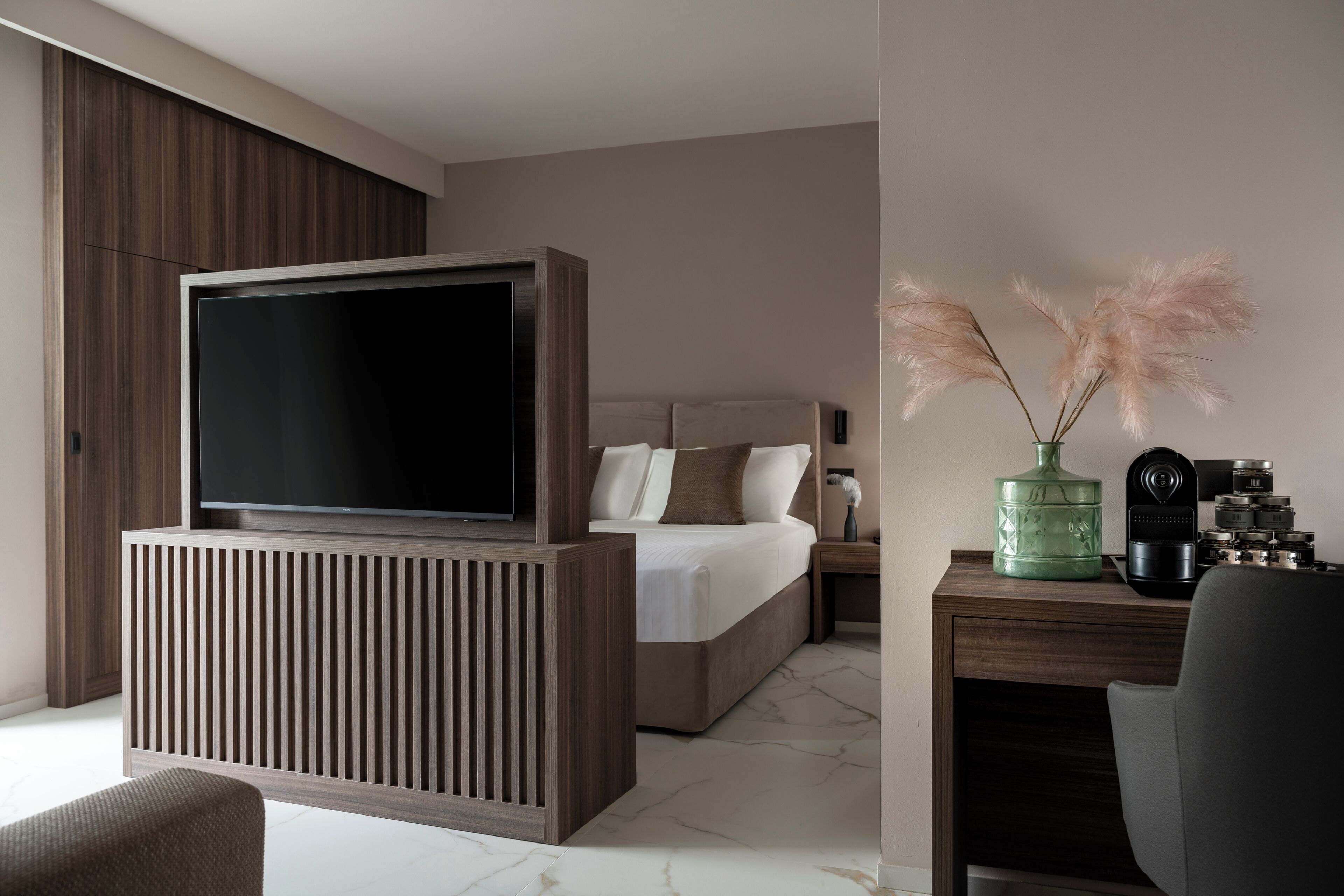Quarto moderno com uma grande televisão de tela plana, cama com roupa de cama branca, cabeceira de madeira, mesa de cabeceira com um vaso de capim-ornamento e uma escrivaninha com uma cadeira.