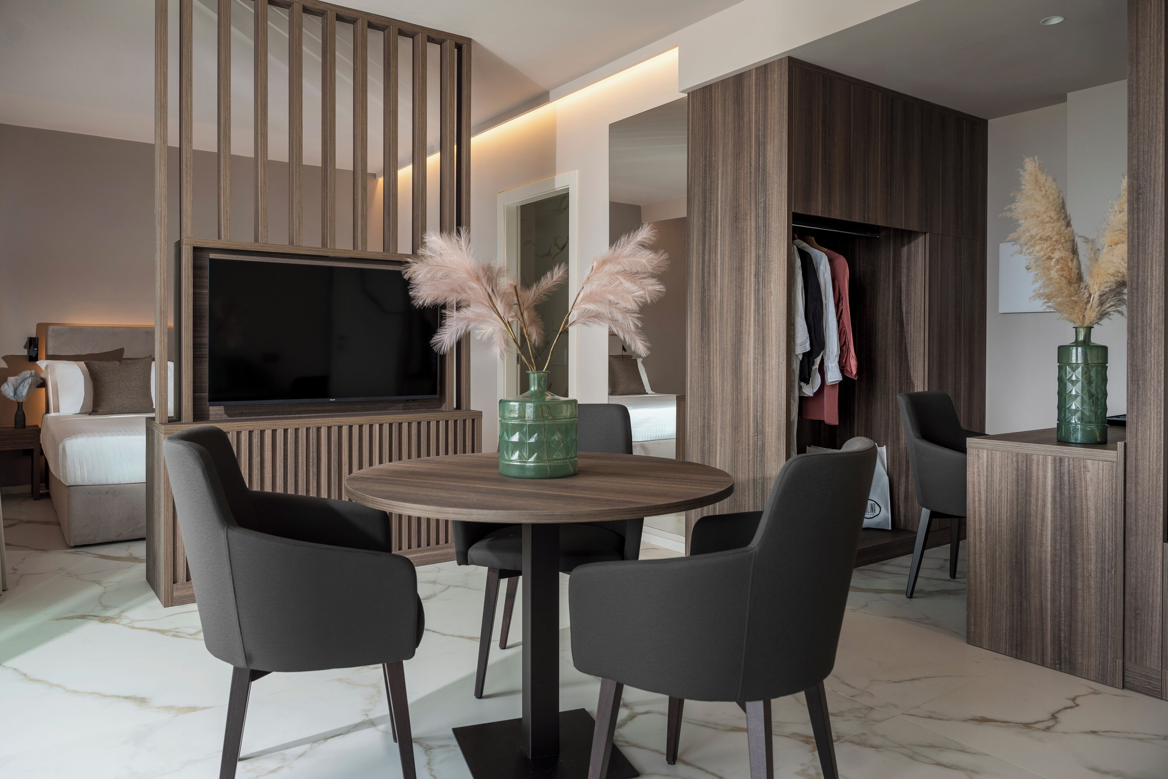 A modern hotel room with a round dining table, two chairs, a TV, a wardrobe, and a green vase with pampas grass.