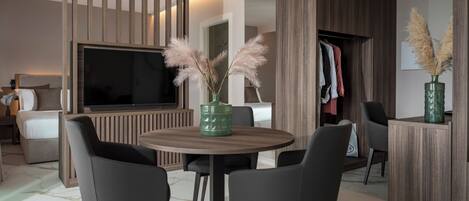 A modern hotel room with a round dining table, two chairs, a TV, a wardrobe, and a green vase with pampas grass.