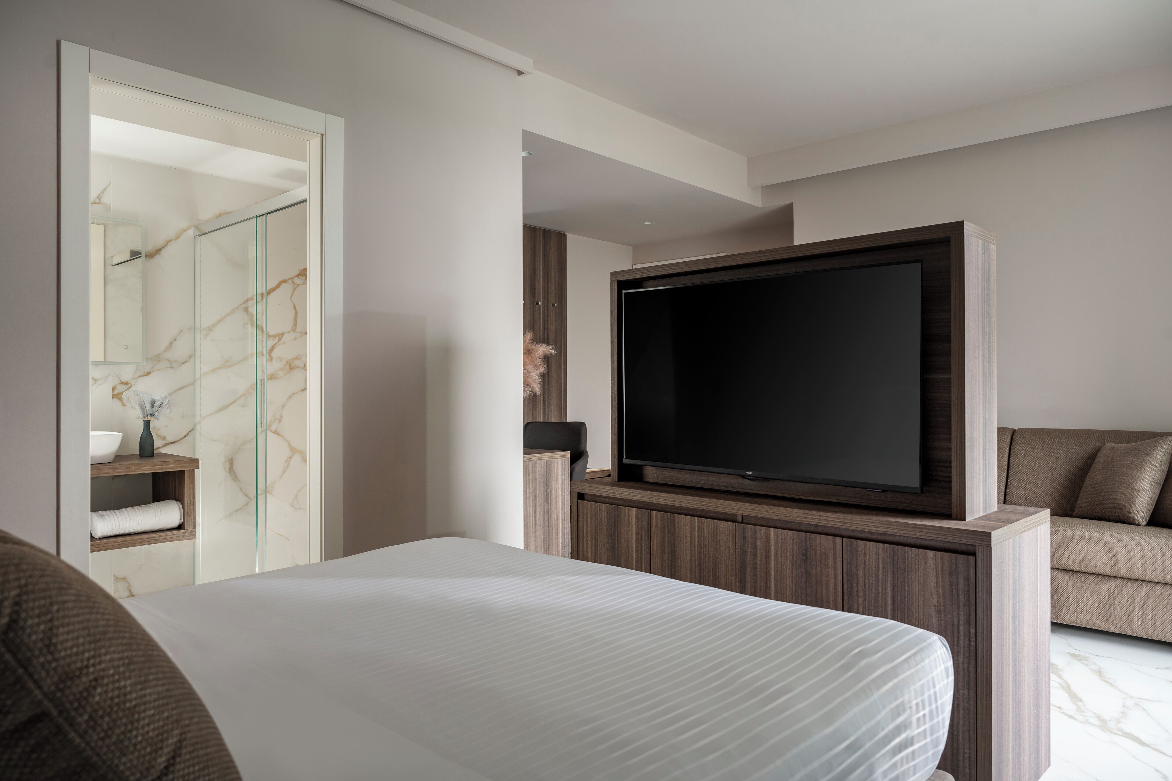 Une chambre d’hôtel moderne dotée d’un grand lit, d’un téléviseur fixé à un meuble en bois, d’une salle de bain en marbre et d’un canapé.