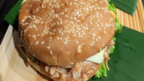 Een sandwich met sesamzaad op een broodje, sla en saus, geserveerd op een houten plank met een groen blad eronder.
