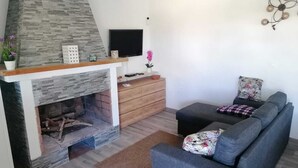 Een woonkamer met een stenen open haard, een flatscreen televisie, een houten kast en een grijze bank met bloemenkussens.