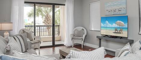 Un salon avec un canapé gris, des fauteuils à rayures, un téléviseur à écran plat et un balcon agrémenté de rideaux blancs.