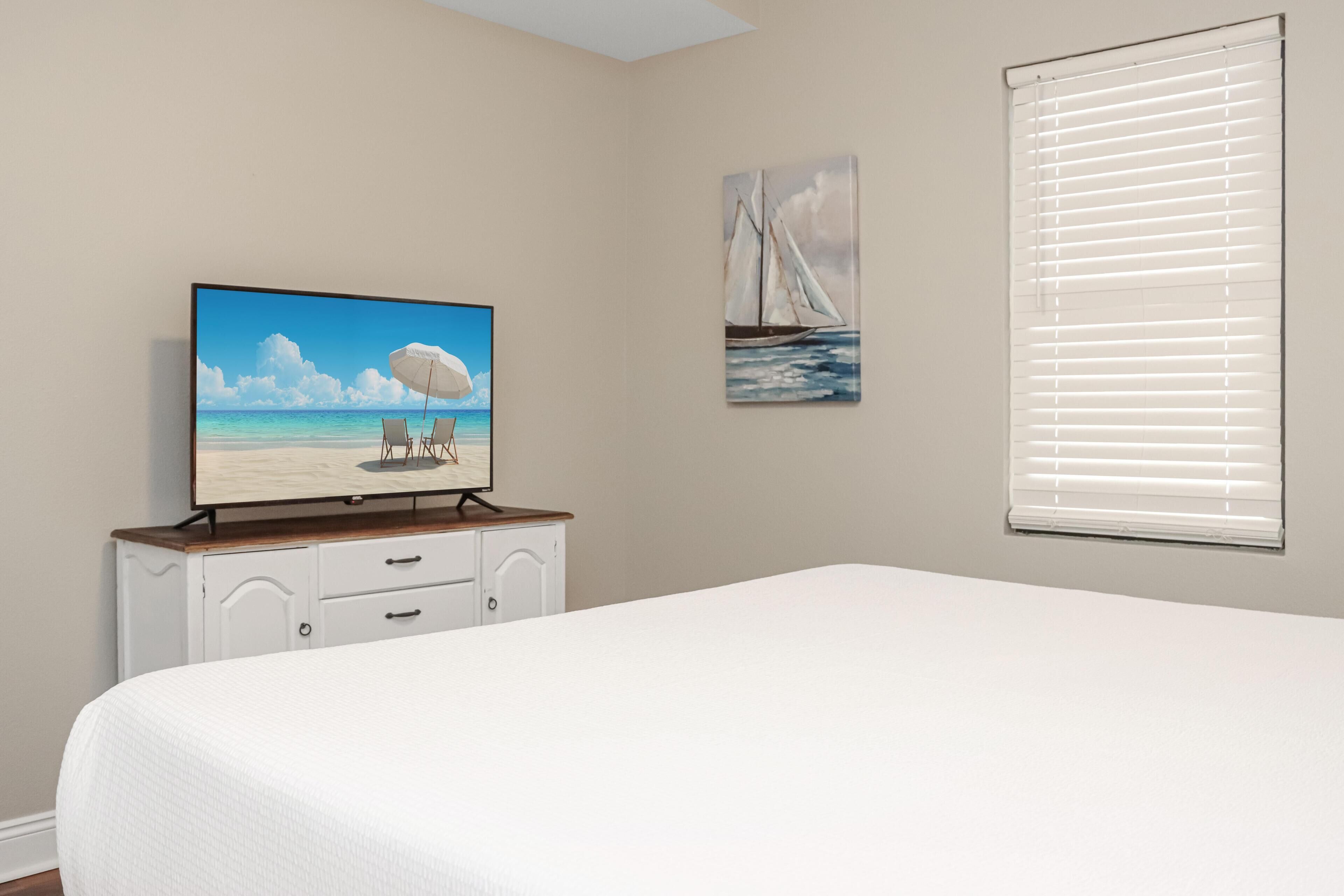 Une chambre à coucher avec un lit, un téléviseur posé sur une commode, une fenêtre avec des stores et deux œuvres d’art accrochées au mur.