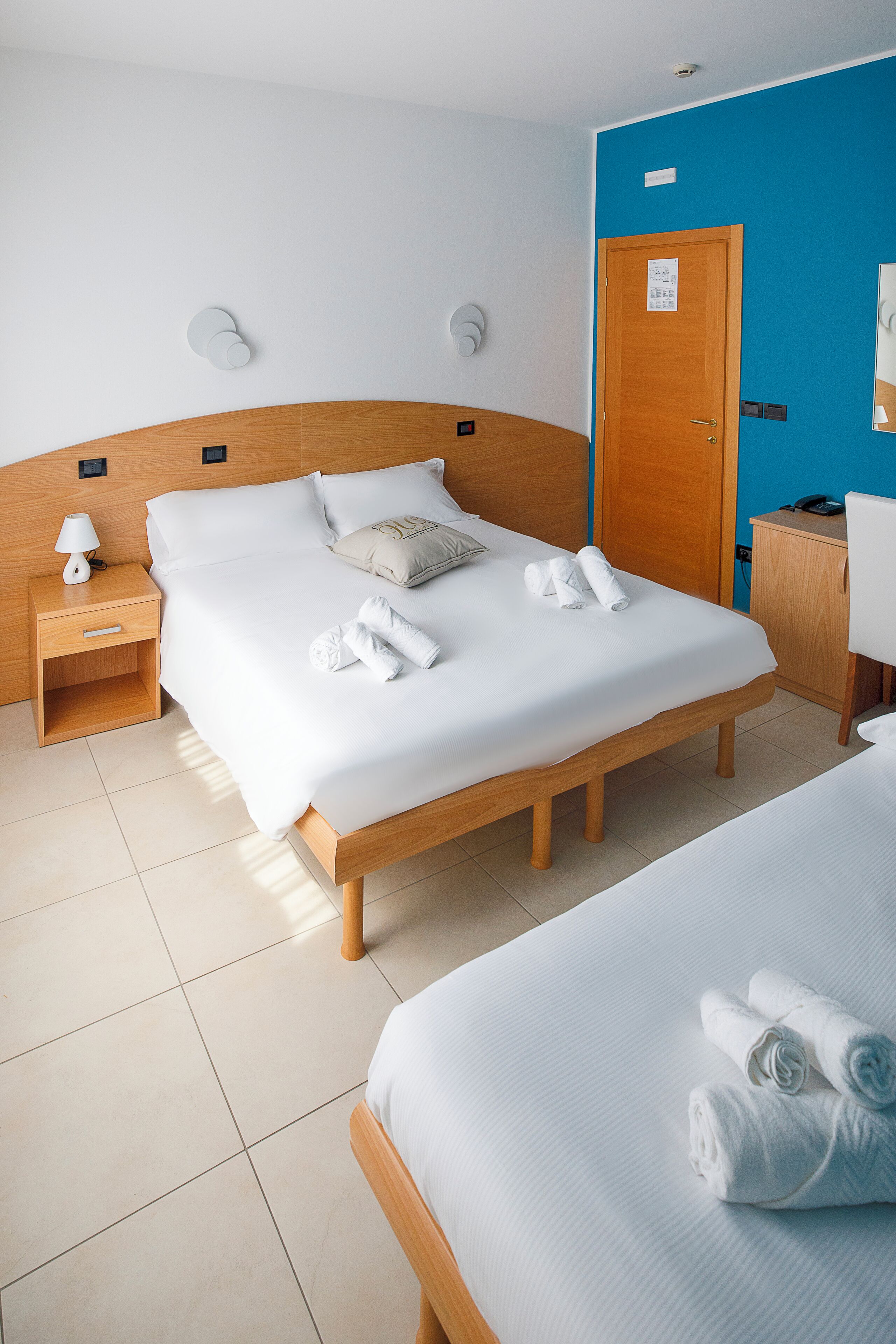 Habitación de hotel con una cama de madera, una mesita de noche con lámpara, un escritorio con silla y pared azul.