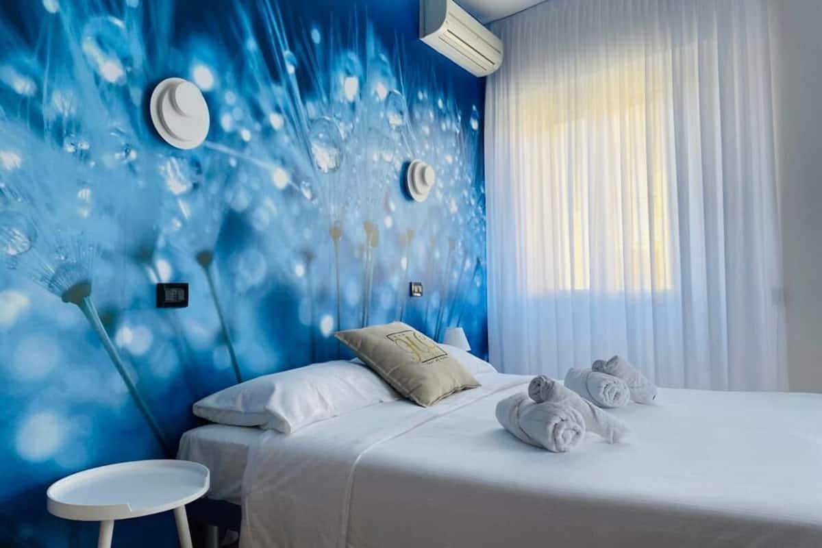 A hotel room with a bed, white bedding, a round table, and a wall with a blue abstract design.