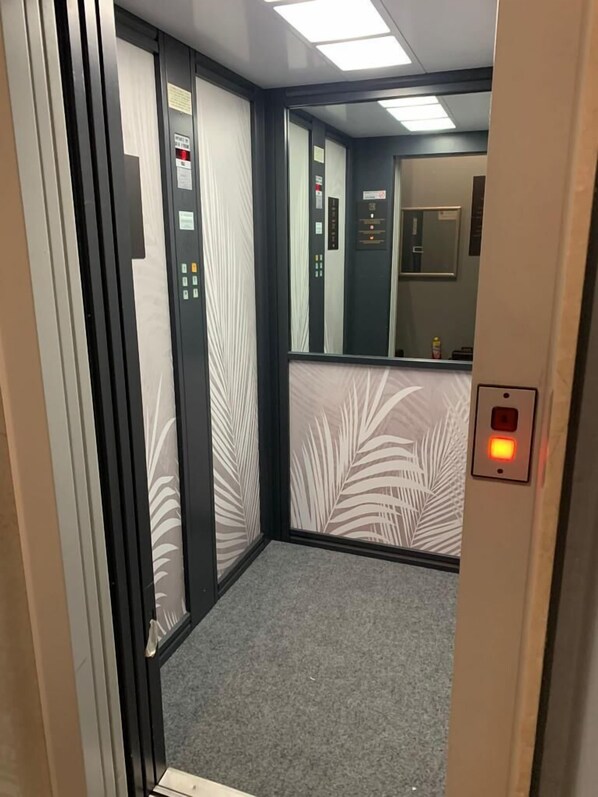 An elevator interior with a grey carpet, a control panel with buttons, and a mirror on the wall. - HC Resort (Lignano Sabbiadoro)