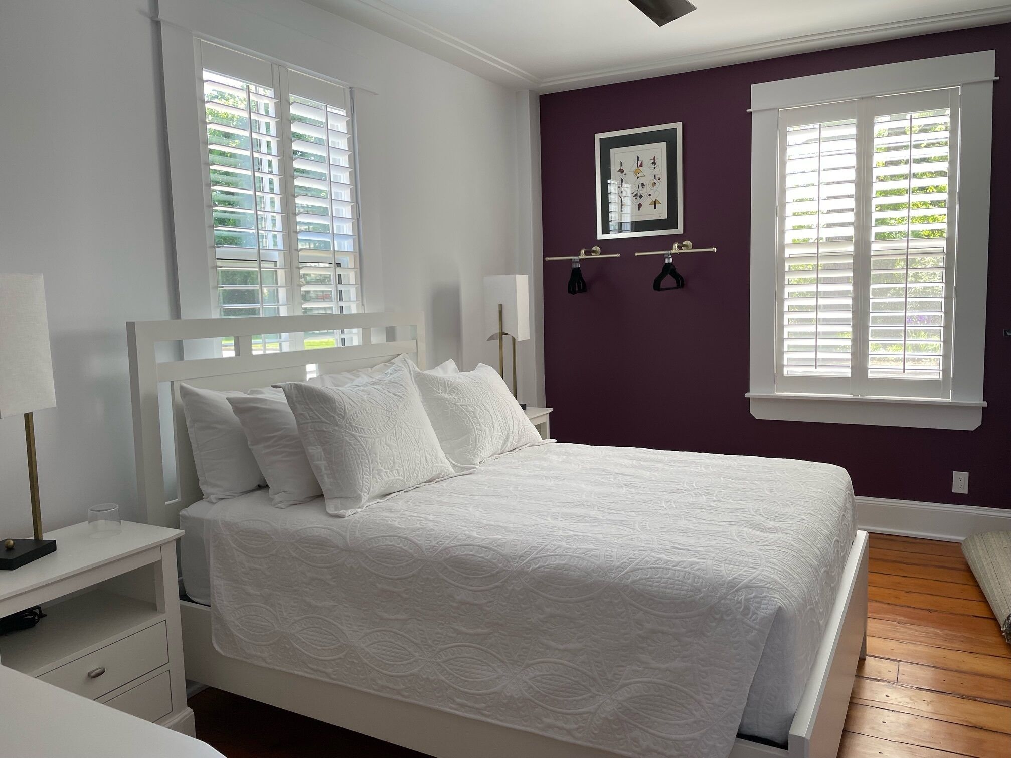Quarto com cama branca, mesa de cabeceira, luminária na parede e janela com persianas.