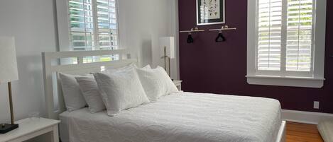 Quarto com cama branca, mesa de cabeceira, luminária na parede e janela com persianas.