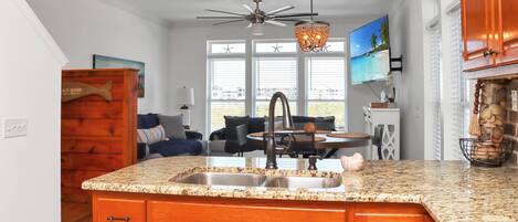 A kitchen with granite countertops, wooden cabinets, a built-in sink, a ceiling fan, and a large window with a view of the beach.
