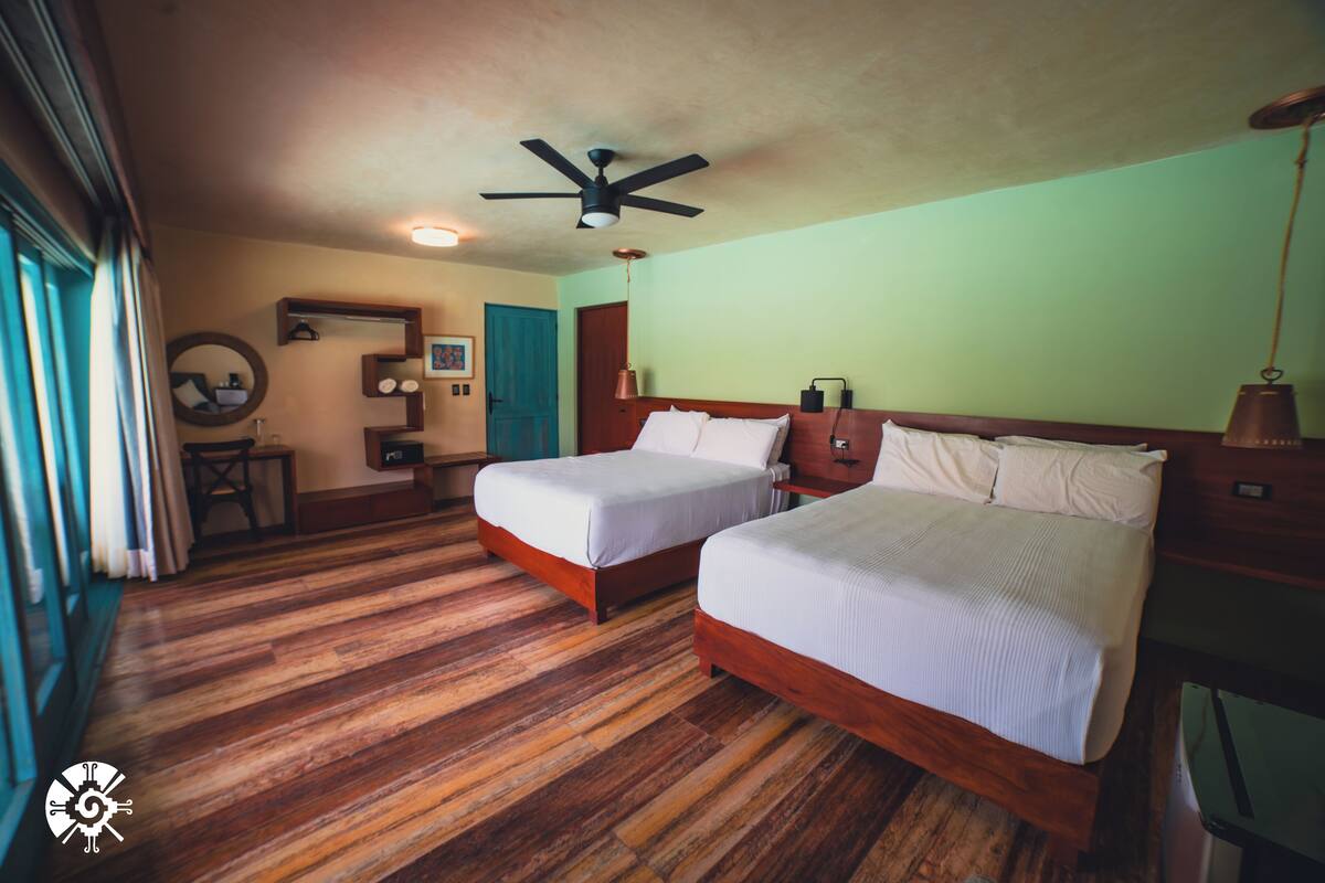 A hotel room with two beds, wooden flooring, a ceiling fan, and a view of the outdoors.