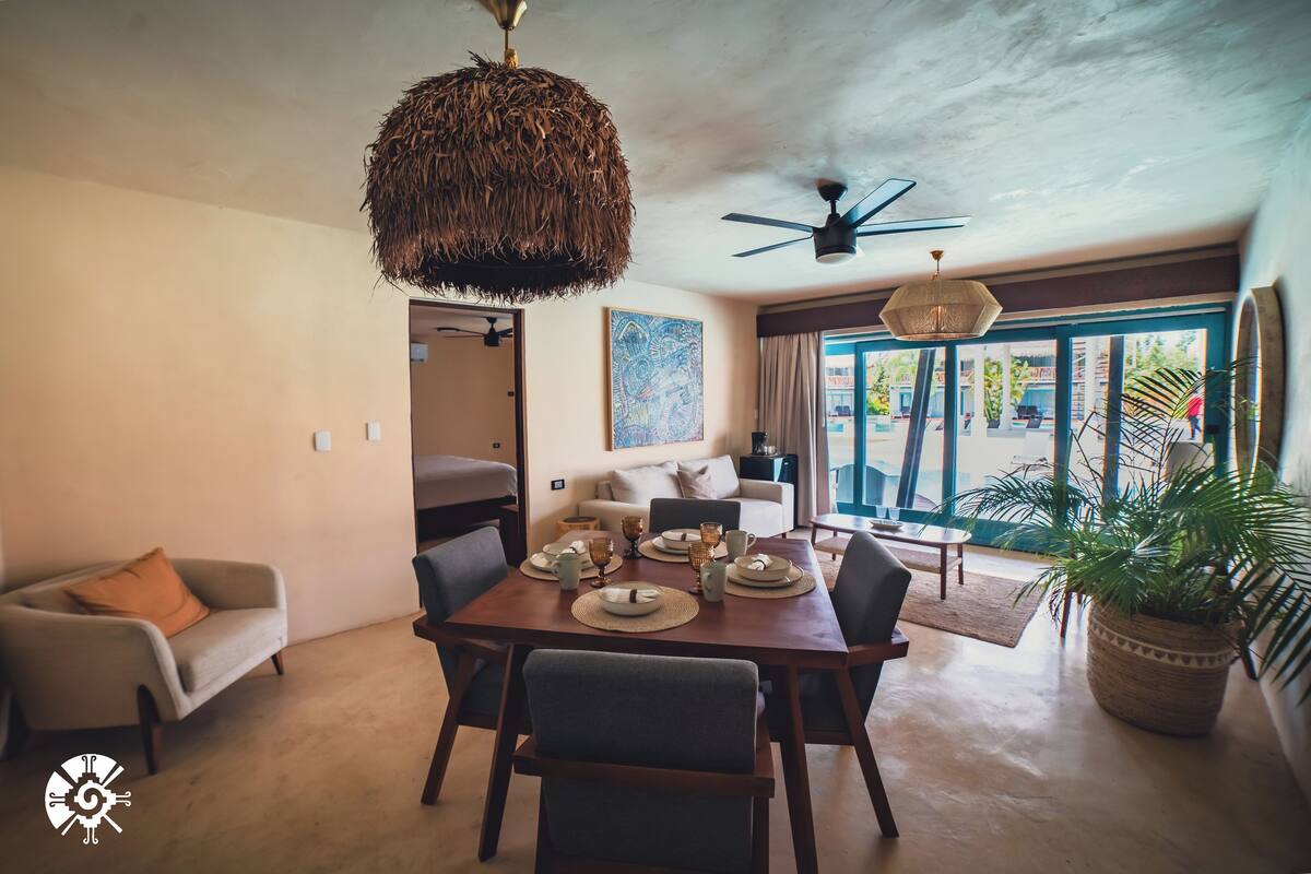A spacious living area with a dining table set for four, a sofa, a ceiling fan, and a large window with a view of the outdoors.