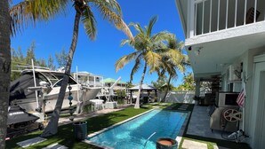 Une piscine bordée de gazon synthétique et de carreaux de pierre, entourée de palmiers et avec un yacht amarré à proximité.