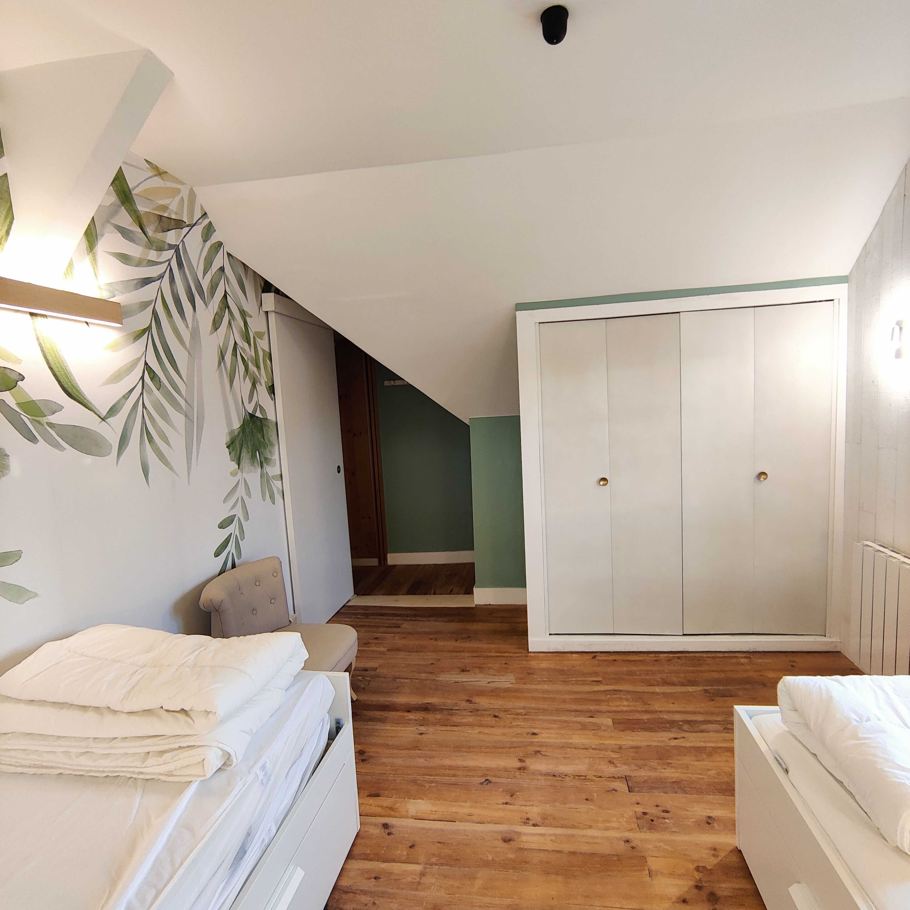 Une chambre avec un plancher en bois, un lit avec une literie blanche, une armoire et un mur orné d’un motif de feuilles.