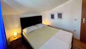 Ein Einzelbett mit dunklem Kopfteil in einem kleinen, schlichten Zimmer mit weißen Wänden und einer geneigten Decke. An der Wand hängen zwei gerahmte Bilder und ein kleiner Nachttisch mit einer Lampe.