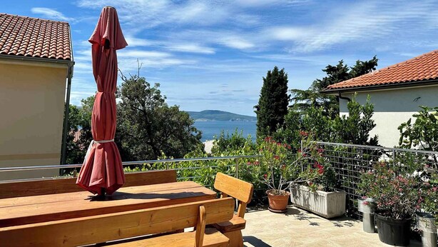 Eine Holzterrasse mit einer Bank und einem roten Sonnenschirm mit Blick auf ein Gewässer und Hügel.