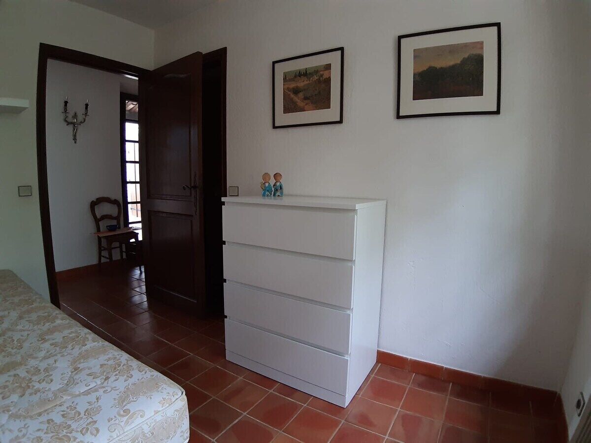 Een slaapkamer met een bed, een witte dressoir, twee ingelijste foto’s aan de muur, en een deur die naar een andere kamer leidt.