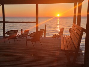 Eine Holzterrasse mit vier Liegestühlen und einer Bank, mit Blick auf einen Sonnenuntergang über dem Wasser.