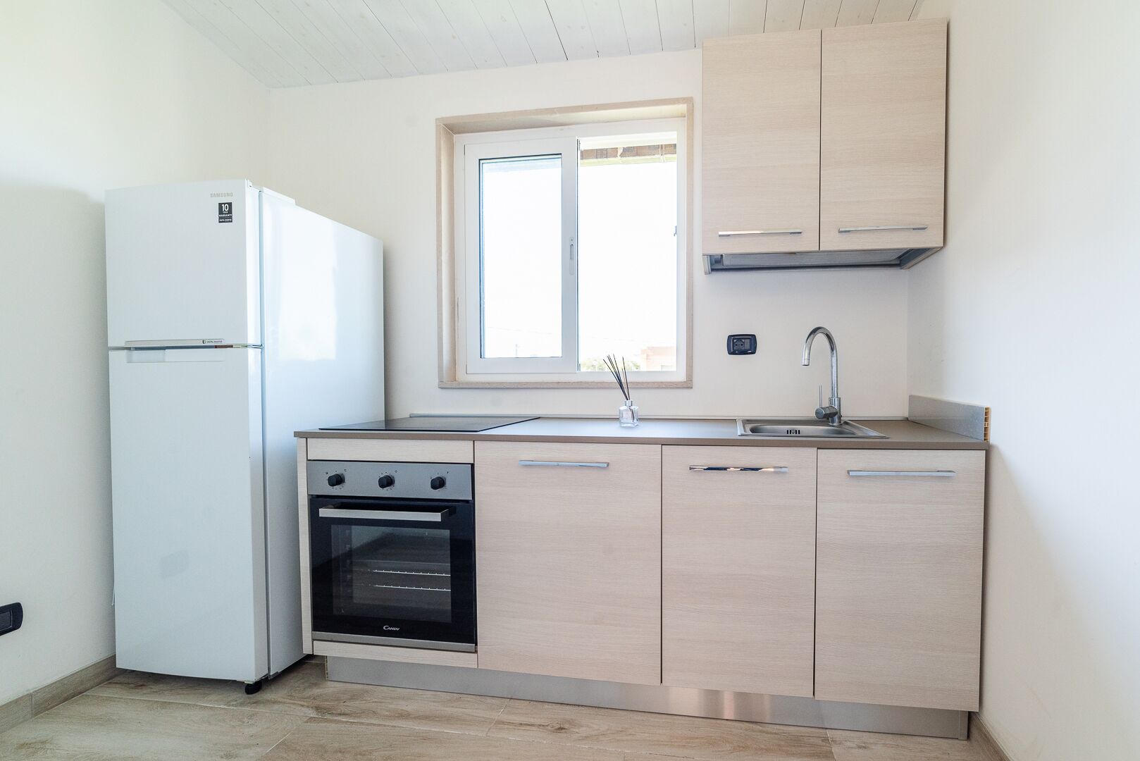 A compact kitchen with a refrigerator, oven, sink, and light wood cabinets.