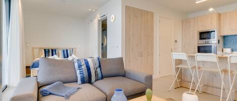 Una sala de estar moderna con un sofá gris, una mesa de comedor de madera y una cocina con sillas blancas y armarios de madera.