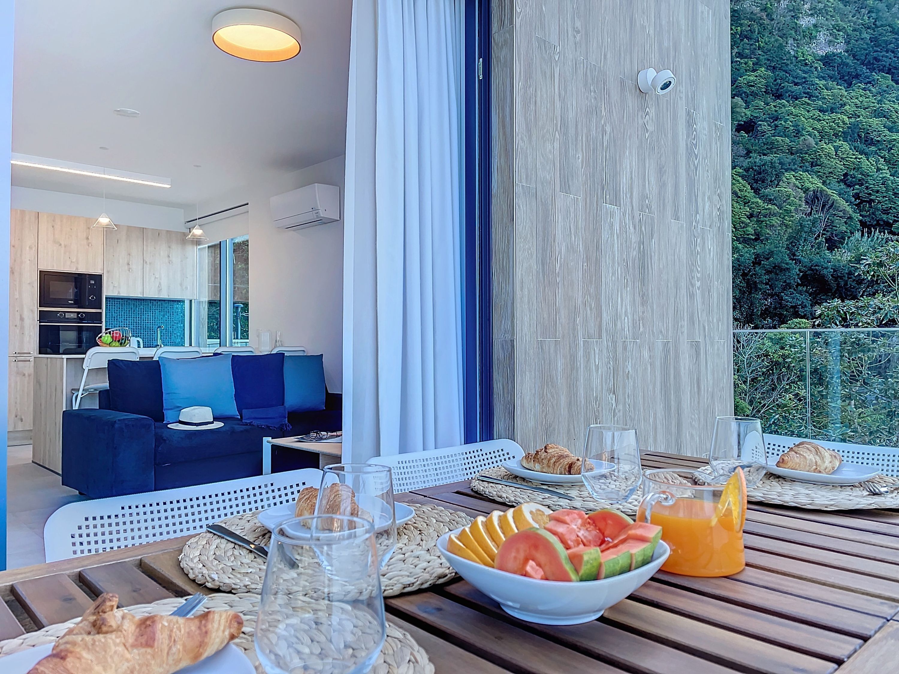 A balcony with breakfast setup including croissants, fruit, and juice, and a view of a modern kitchen and living area.