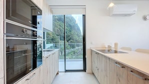 A modern kitchen with wooden cabinets, a built-in oven, and a large sliding glass door leading to a balcony with a view of greenery. - Mar Yam View by Atlantic Holiday (Santana)