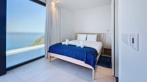 A bedroom with a large window overlooking the sea, a bed with a blue bedspread, and a wooden headboard. - Mar Yam View by Atlantic Holiday (Santana)