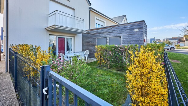A modern residential building with a balcony, a small garden with yellow flowering plants, and a paved driveway. - Les Anémones de Langrune - Avec jardin clos (Langrune-sur-Mer)
